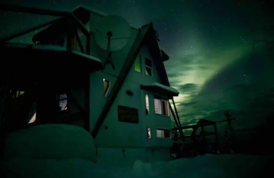 Nighttime view of a cozy treehouse structure under glowing green northern lights in Fairbanks, Alaska