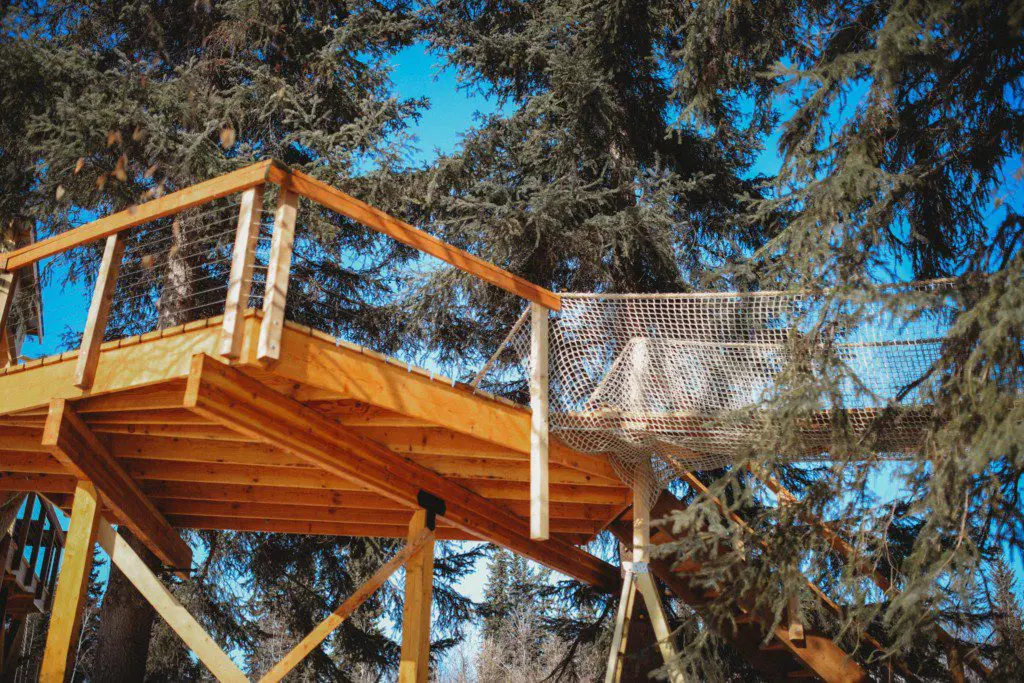 Raised wooden treehouse deck with rope netting nestled in tall pine trees in Fairbanks, Alaska