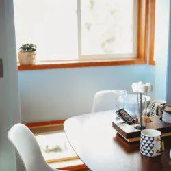 Small dining table with mugs, books, and white chairs by a sunny window in a Fairbanks Alaska treehouse