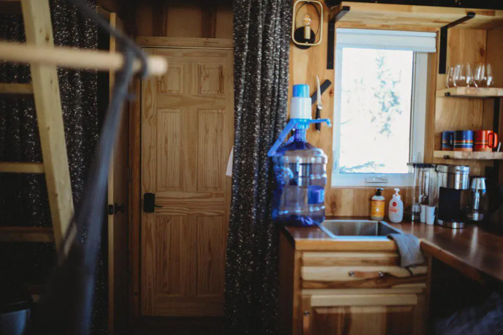 Small wood-paneled kitchen with water jug, sink, and window in a Fairbanks Alaska treehouse rental