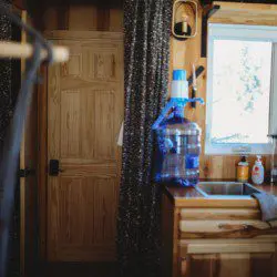 Small wood-paneled kitchen with water jug, sink, and window in a Fairbanks Alaska treehouse rental