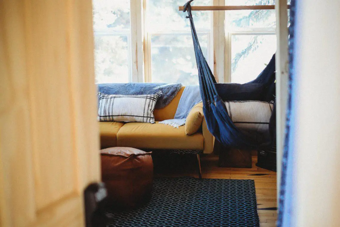Interior view of a sunlit room with hammock chair, sofa, and pouf in a Fairbanks Alaska treehouse rental
