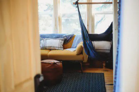 Interior view of a sunlit room with hammock chair, sofa, and pouf in a Fairbanks Alaska treehouse rental