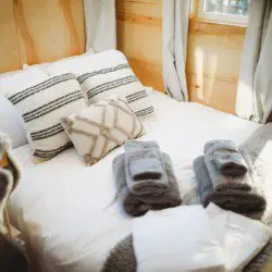 Made bed with soft pillows, gray towels, and sunlight streaming through windows in Fairbanks treehouse
