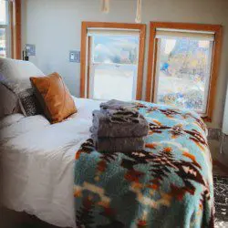 Cozy bed with folded towels and colorful blanket near large windows in a Fairbanks Alaska treehouse