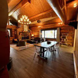 Large wood-beamed lodge interior with chandelier, dining table, seating area, and fireplace in Fairbanks, Alaska