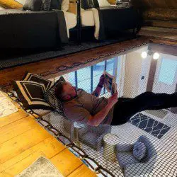 Person reading while lying in a netted hammock loft above a cozy living area in a Fairbanks Alaska treehouse