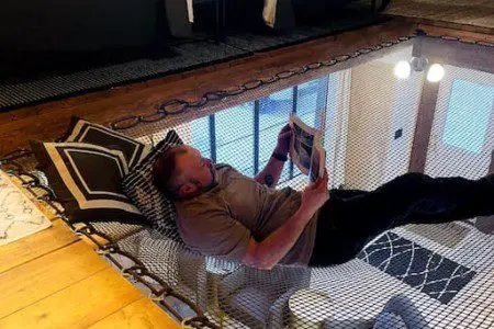 Person reading while lying in a netted hammock loft above a cozy living area in a Fairbanks Alaska treehouse
