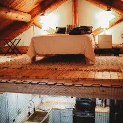 Bed in a wood-beamed loft above kitchen area in a rustic Fairbanks Alaska treehouse rental