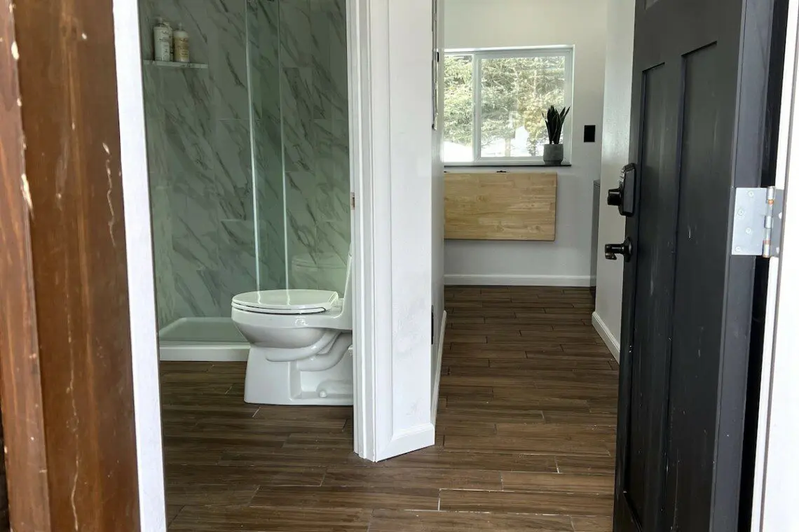 Open black door revealing a clean bathroom with glass shower and wood-look tile flooring in a Fairbanks rental
