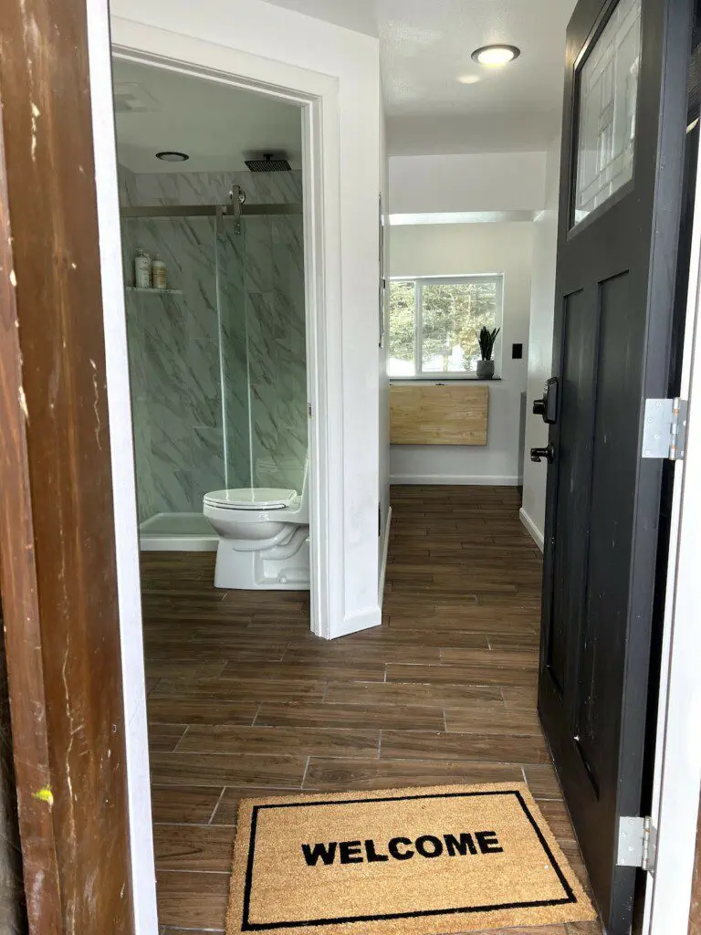 Open black door revealing a clean bathroom with glass shower and wood-look tile flooring in a Fairbanks rental