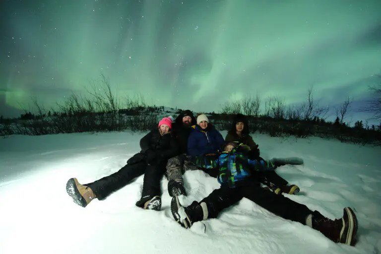 Family sitting in snow under vibrant northern lights in Fairbanks during a winter night