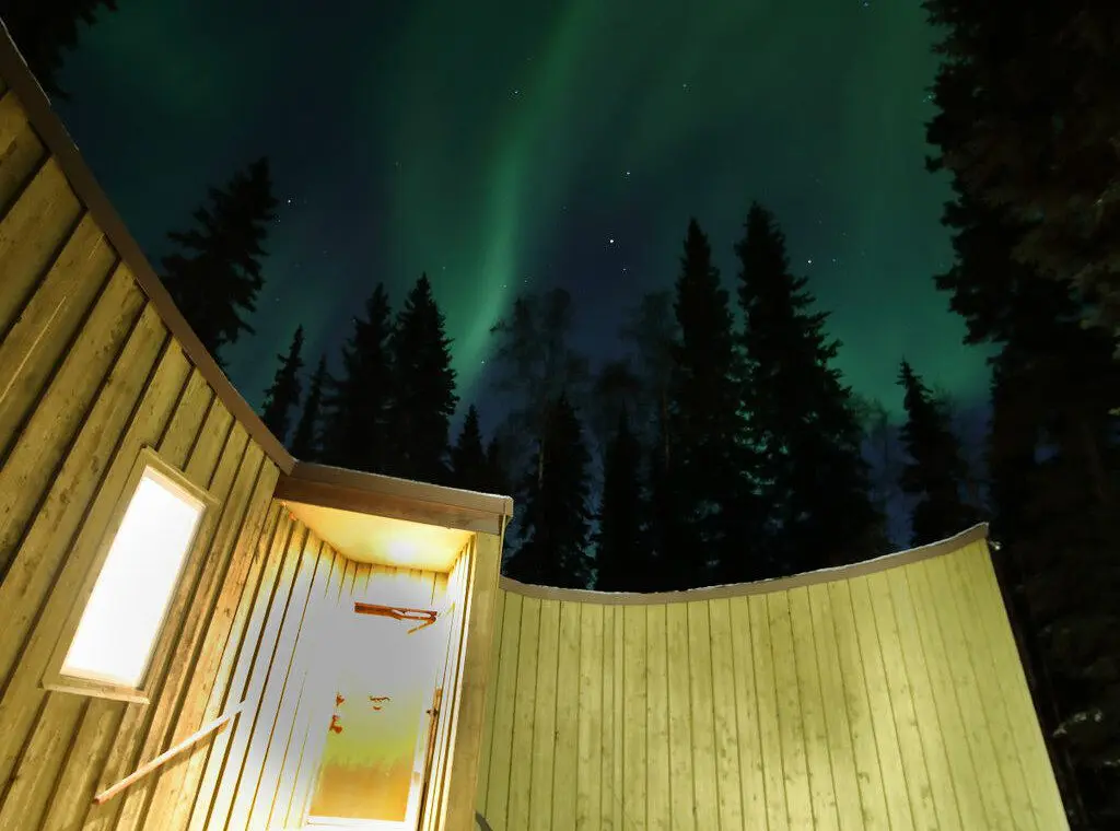 Glowing cabin entrance under vibrant northern lights and pine trees in Fairbanks, Alaska