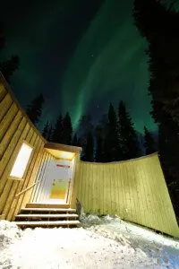 Glowing cabin entrance under vibrant northern lights and pine trees in Fairbanks, Alaska