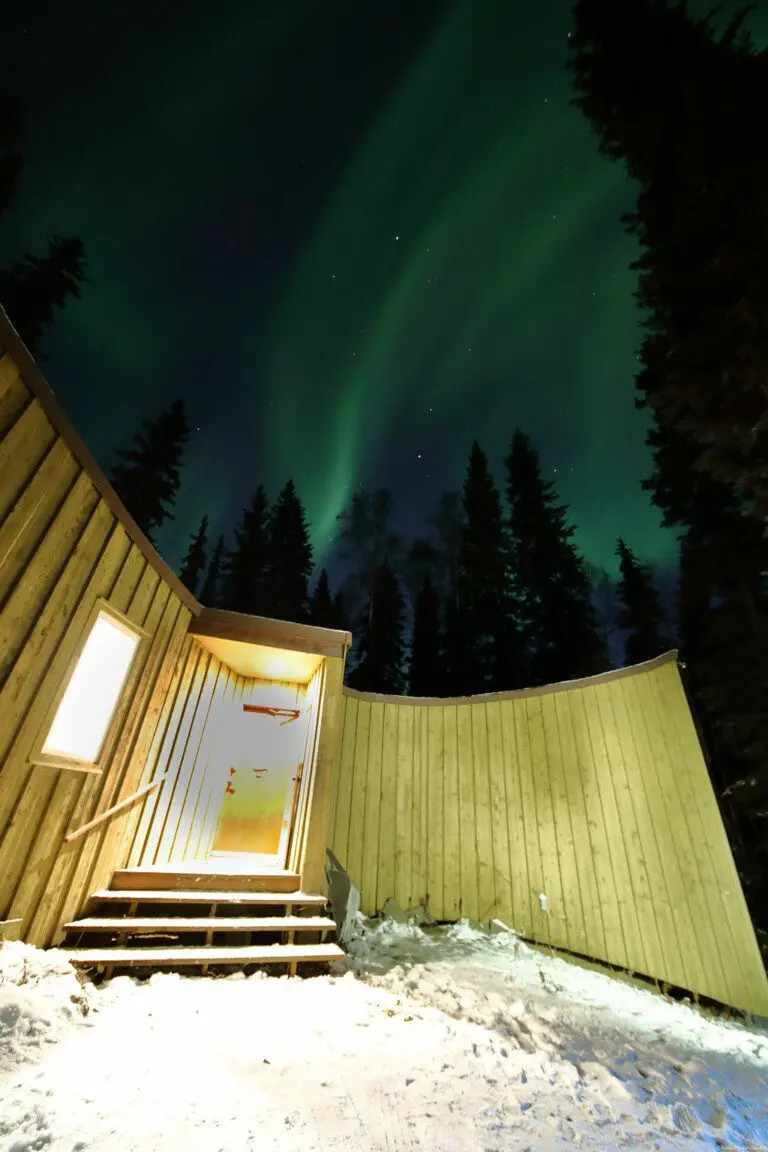 Glowing cabin entrance under vibrant northern lights and pine trees in Fairbanks, Alaska