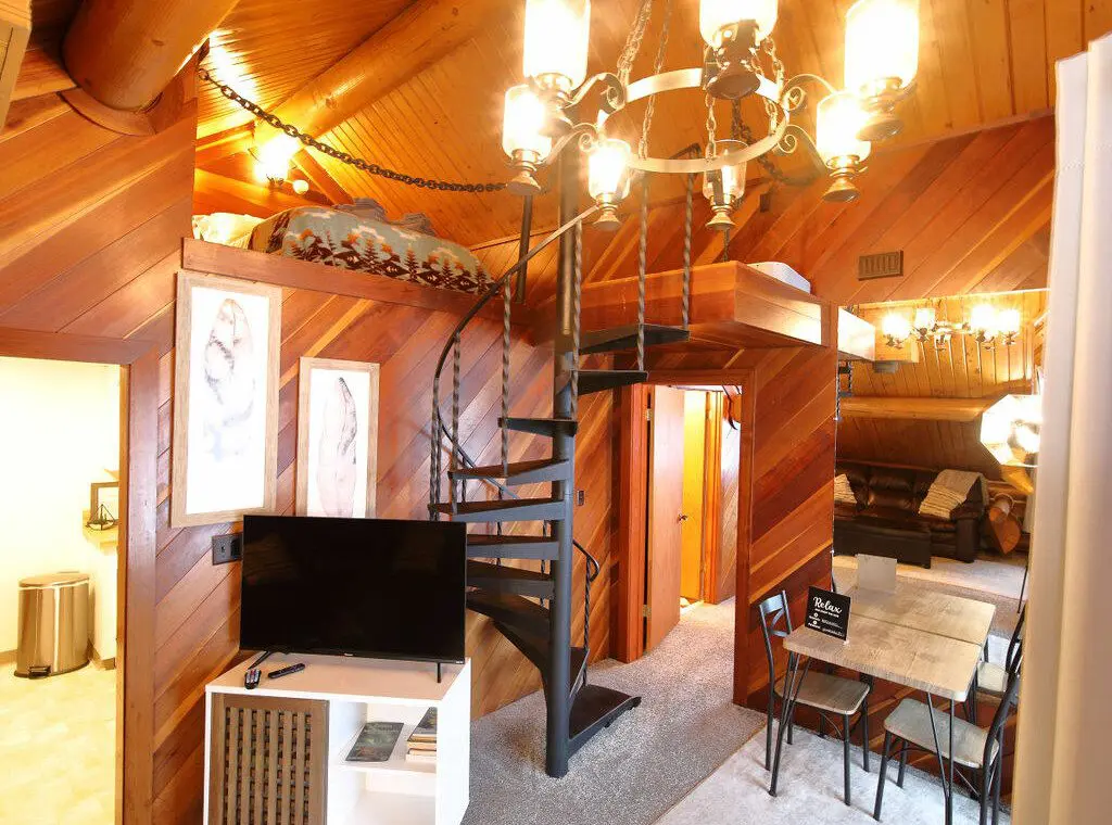 Interior view of a log cabin in Fairbanks with a black spiral staircase, chandelier, TV stand, and rustic wood accents.