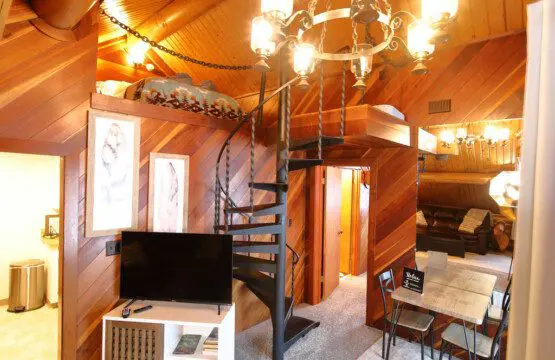 Interior view of a log cabin in Fairbanks with a black spiral staircase, chandelier, TV stand, and rustic wood accents.