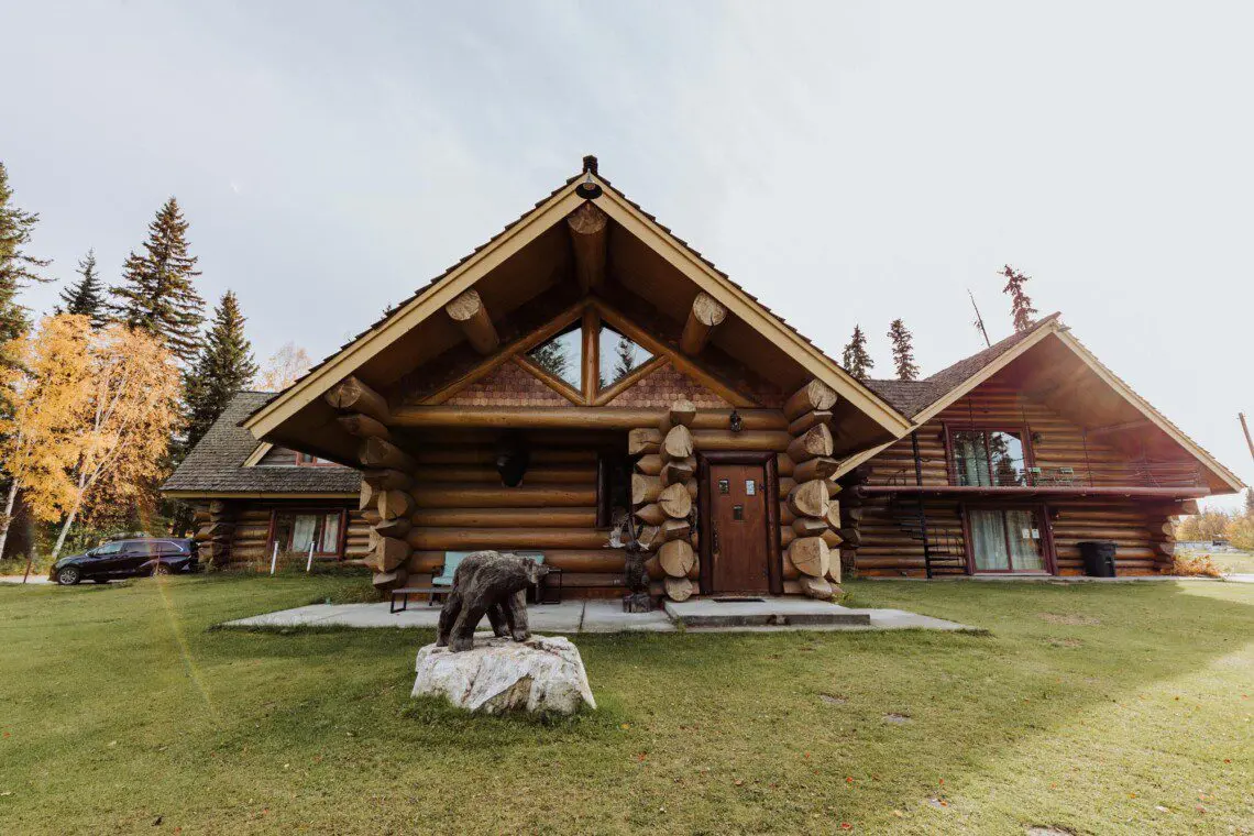 Front view of a log cabin lodge with carved bear sculpture and green lawn at Alaska Adventure Lodge in Fairbanks, Alaska