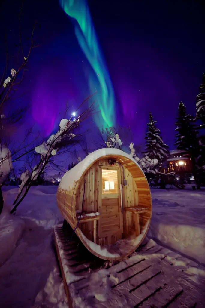 Snow-covered barrel sauna glowing under northern lights in Fairbanks Alaska near treehouse rentals