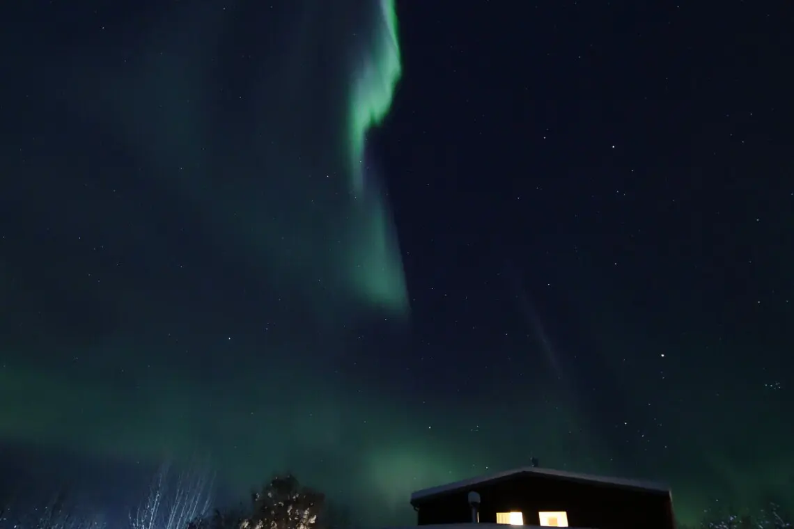 Glowing northern lights above snowy Fairbanks treehouse lodge at night with warm interior lighting