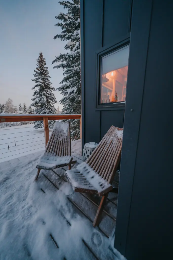 Two wooden chairs on a snow-covered balcony outside a modern treehouse rental in Fairbanks Alaska