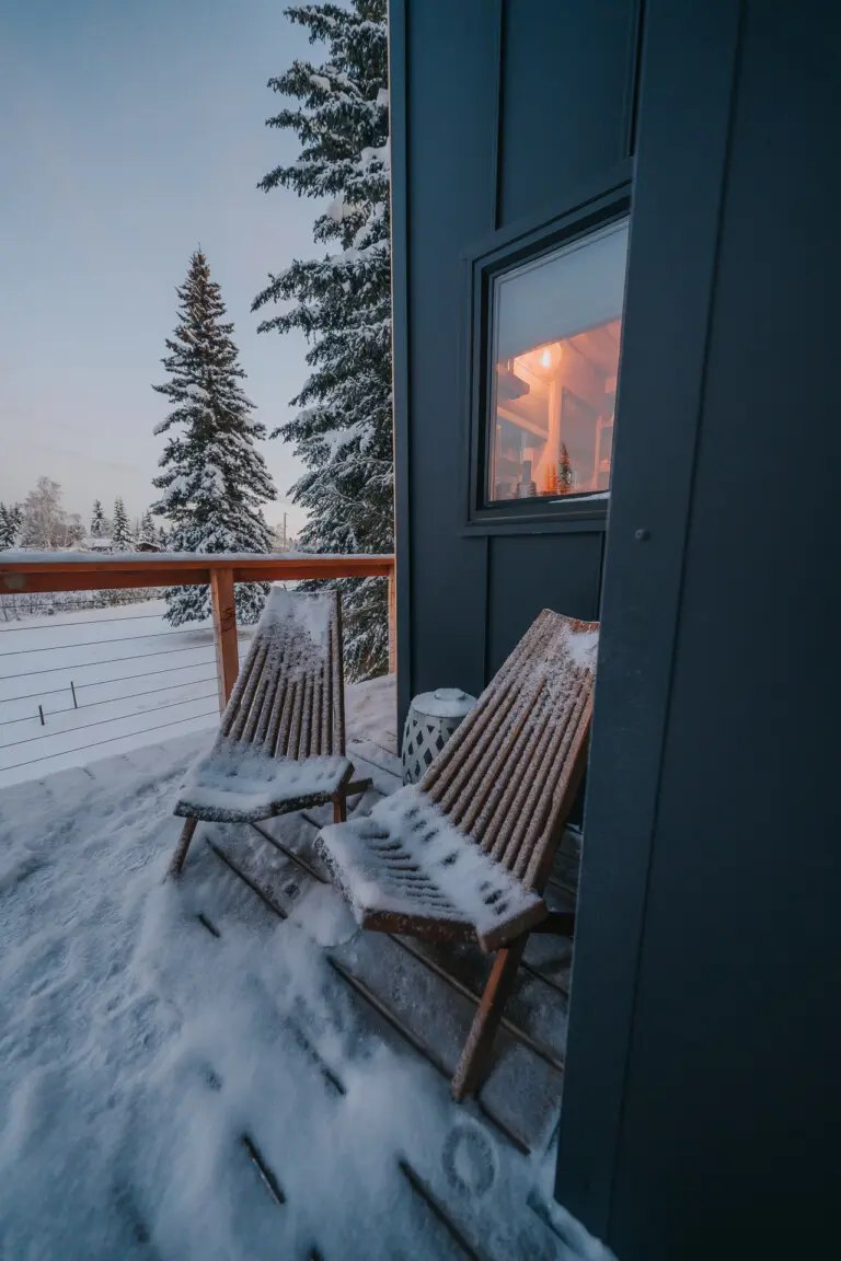Two wooden chairs on a snow-covered balcony outside a modern treehouse rental in Fairbanks Alaska