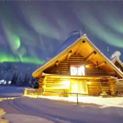 Log cabin in snowy Fairbanks lit under green aurora at an Alaska adventure lodge.