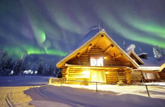 Log cabin in snowy Fairbanks lit under green aurora at an Alaska adventure lodge.