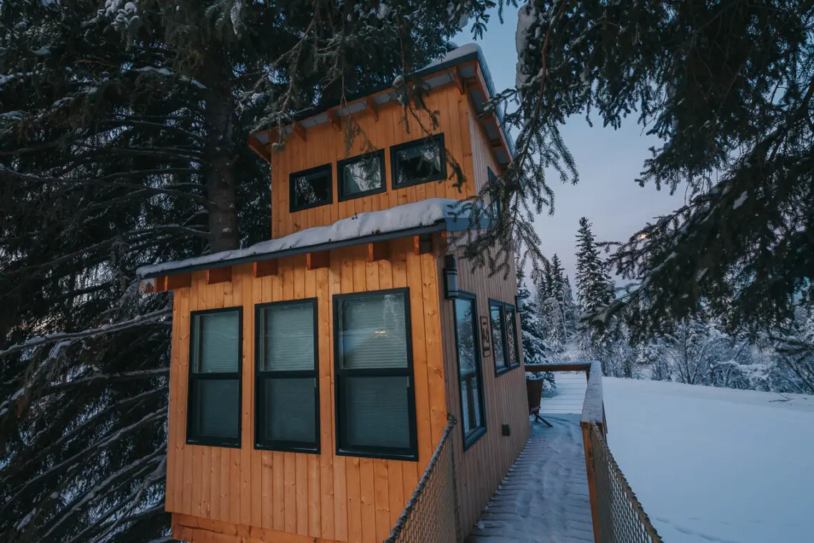 Couple standing on a snowy wooden treehouse deck surrounded by pine trees in Fairbanks Alaska