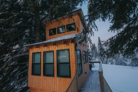 Couple standing on a snowy wooden treehouse deck surrounded by pine trees in Fairbanks Alaska