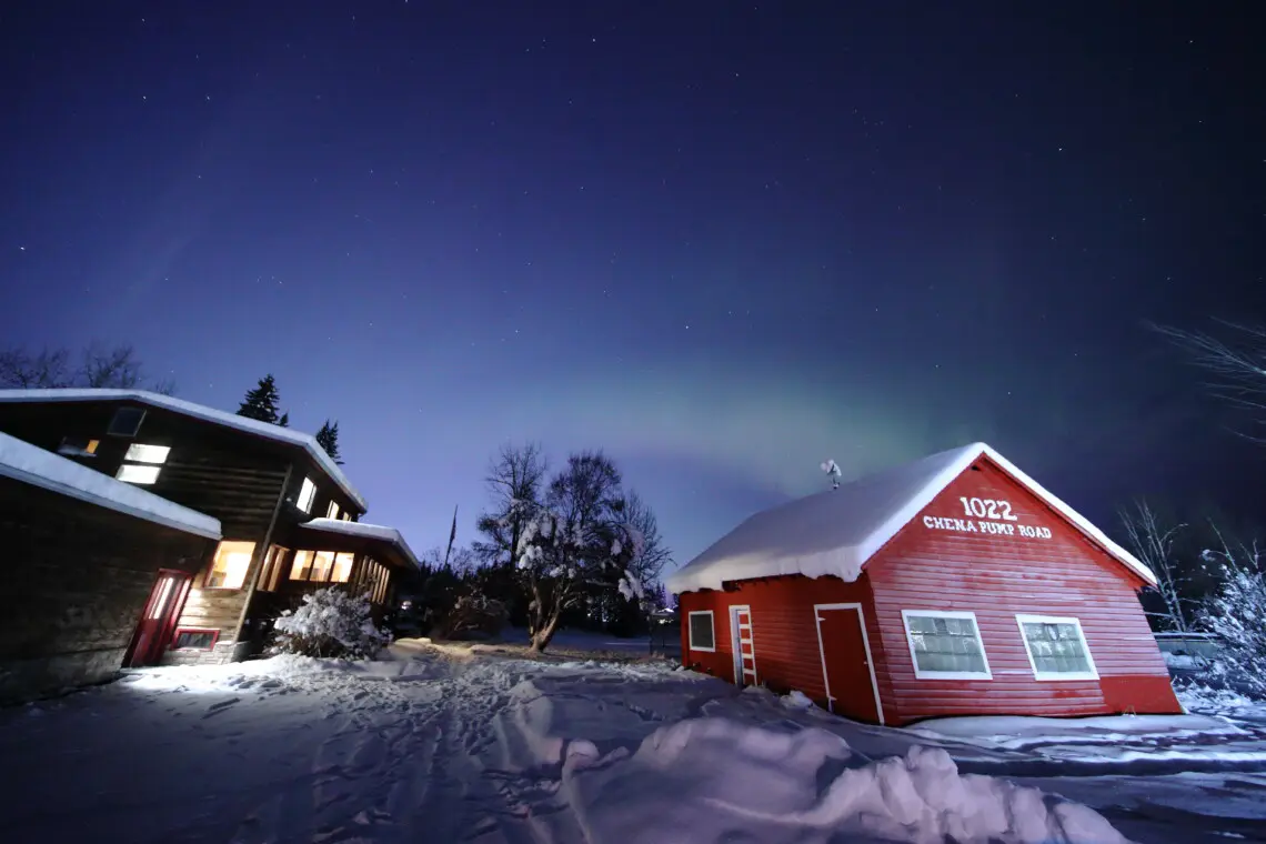 Rustic lodge and red cabin under aurora lights on Chena Pump Road in Fairbanks, Alaska.