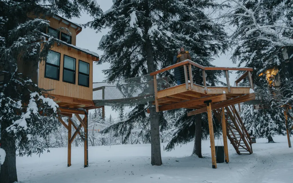 Couple standing on a snowy wooden treehouse deck surrounded by pine trees in Fairbanks Alaska