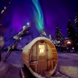 Northern lights over barrel sauna at Alaska adventure lodge in Fairbanks – unique treehouse-style winter stay.