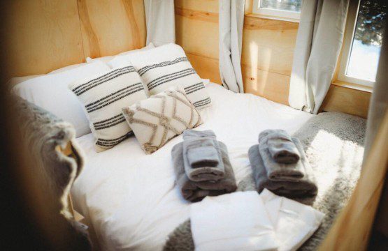 Made bed with soft pillows, gray towels, and sunlight streaming through windows in Fairbanks treehouse