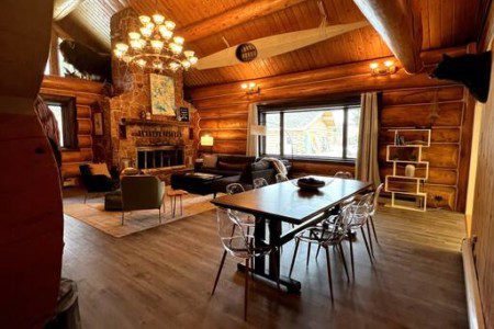 Large wood-beamed lodge interior with chandelier, dining table, seating area, and fireplace in Fairbanks, Alaska