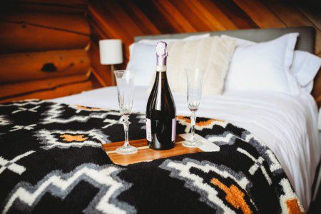 Bottle of champagne and glasses on bed with patterned blanket in cozy Fairbanks treehouse rental