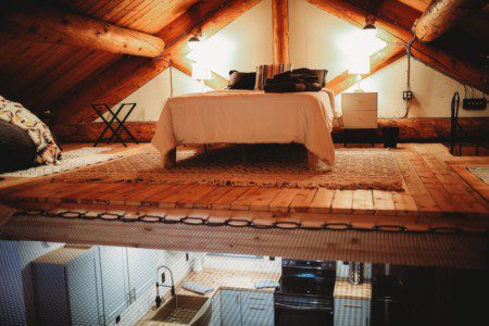 Bed in a wood-beamed loft above kitchen area in a rustic Fairbanks Alaska treehouse rental