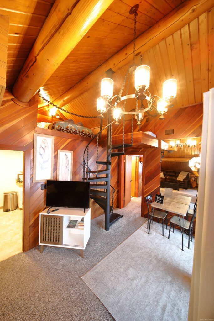 Interior view of a log cabin in Fairbanks with a black spiral staircase, chandelier, TV stand, and rustic wood accents.