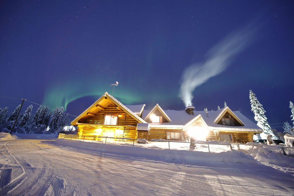 Illuminated log cabin lodge with smoke rising and northern lights overhead in snowy Fairbanks Alaska