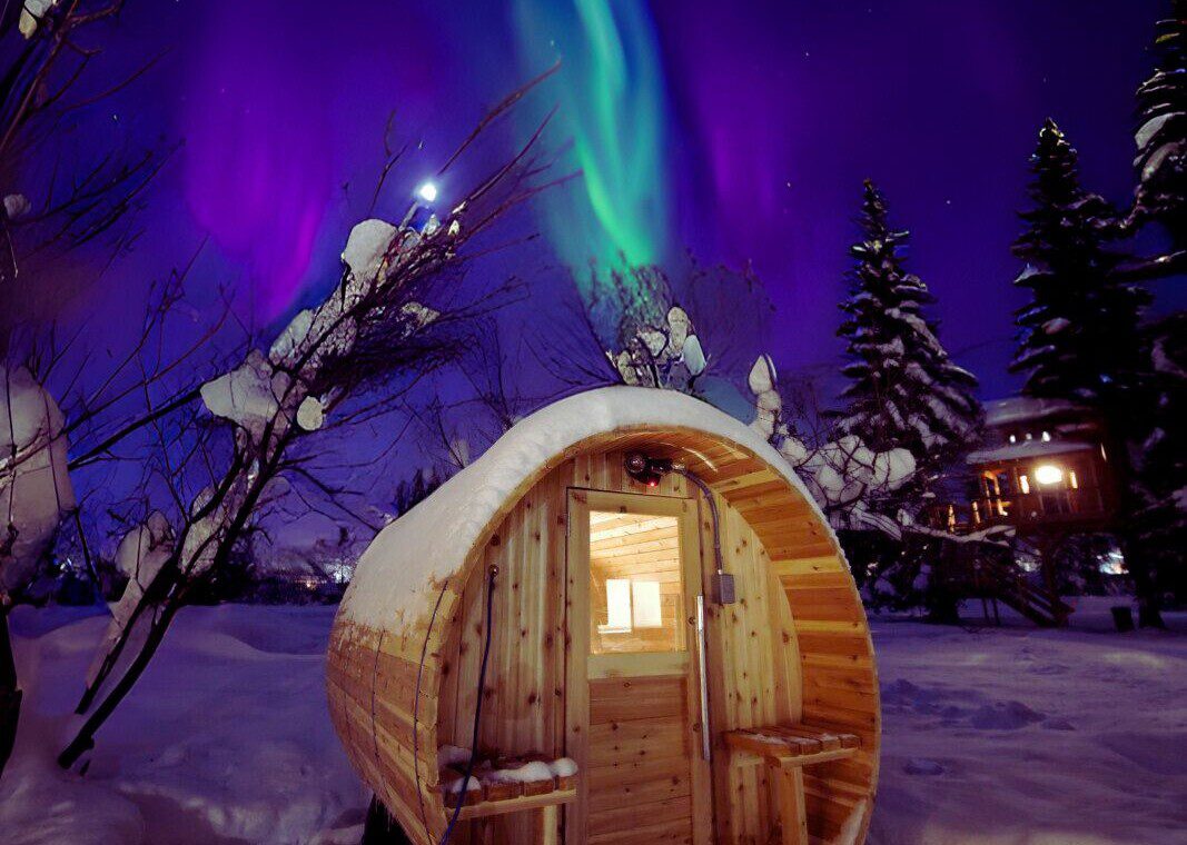 Snow-covered barrel sauna glowing under northern lights in Fairbanks Alaska near treehouse rentals