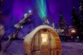 Snow-covered barrel sauna glowing under northern lights in Fairbanks Alaska near treehouse rentals