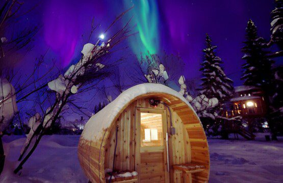 Snow-covered barrel sauna glowing under northern lights in Fairbanks Alaska near treehouse rentals