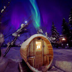 Barrel sauna in snowy Fairbanks with aurora above at Alaska adventure lodge.