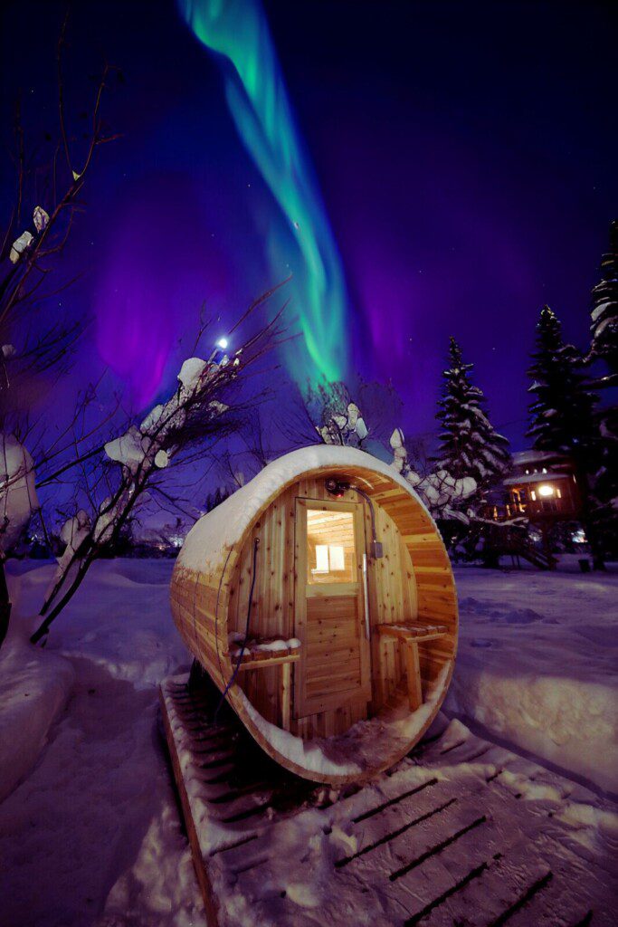 Barrel sauna in snowy Fairbanks with aurora above at Alaska adventure lodge.