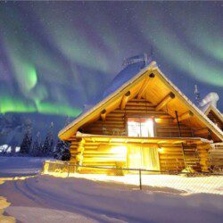 Log cabin in snowy Fairbanks lit under green aurora at an Alaska adventure lodge.