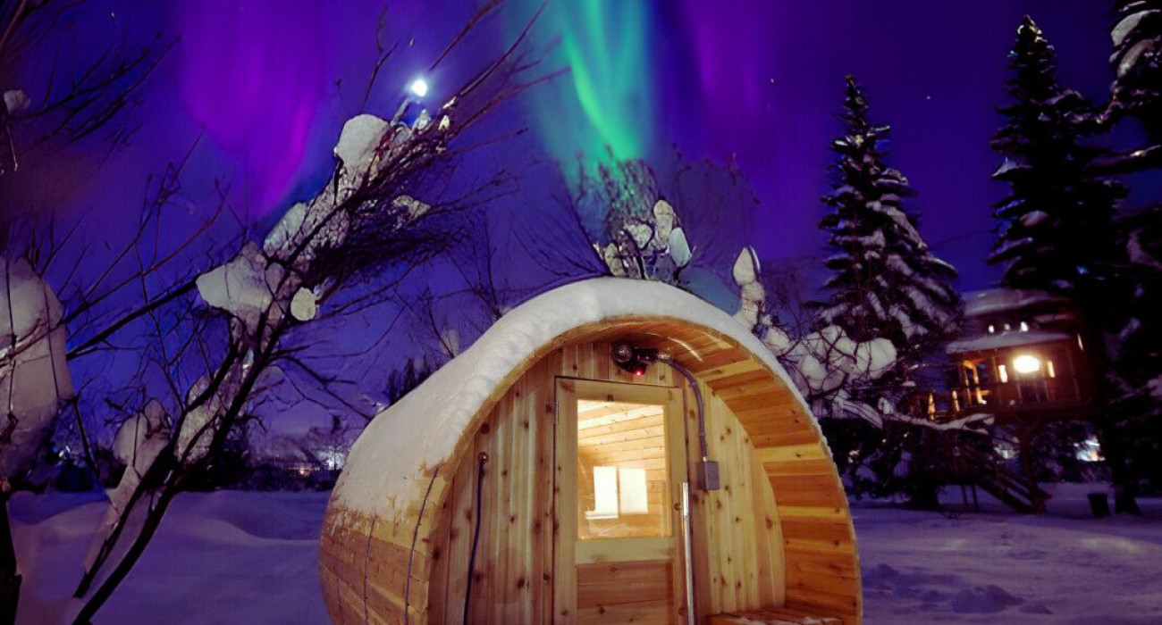 Snow-covered barrel sauna glowing under northern lights in Fairbanks Alaska near treehouse rentals