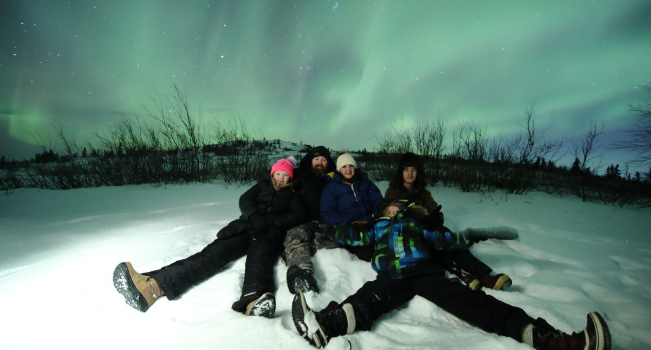Family sitting in snow under vibrant northern lights in Fairbanks during a winter night